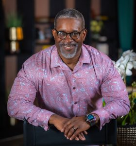 Portrait of Pastor Taiwan Simmons wearing a patterned shirt, smiling confidently while seated in a professional setting